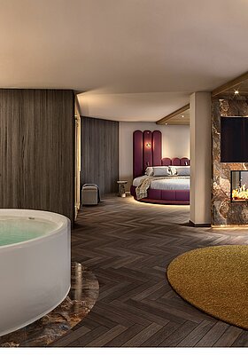 Visualisierung einer luxuriösen Hotel-Wellness-Suite. Im Vordergrund steht eine große, freistehende Whirlpool-Badewanne auf einem marmorierten Sockel. Rechts daneben befindet sich ein gemütlicher Sitzbereich mit einem runden Loungesessel, einem Beistelltisch und einem Kamin unter einem Fernseher. Im Hintergrund ist ein großer kreisförmiger Schlafbereich mit einem markanten violetten Kopfteil zu sehen. Die Suite zeichnet sich durch dunkles Fischgrätparkett, strukturierte Holz- und Marmorwände sowie warme, gedimmte Beleuchtung aus.